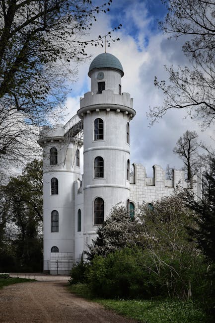 A white, fairy-tale style castle tower featuring rounded turrets and narrow, arched windows, set amidst a lush garden landscape. The surrounding foliage includes tall trees with budding leaves and den