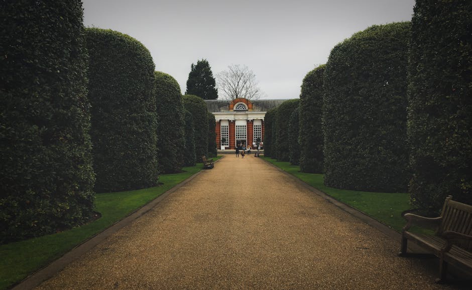 A white, fairy-tale style castle tower featuring rounded turrets and narrow, arched windows, set amidst a lush garden landscape. The surrounding foliage includes tall trees with budding leaves and den