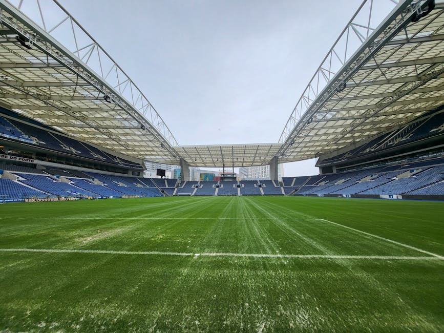 A wide view of a sports stadium featuring a well-maintained, lush green playing field with freshly mowed grass and visible mowing stripes. Surrounding the field are tiered seating areas with blue and 