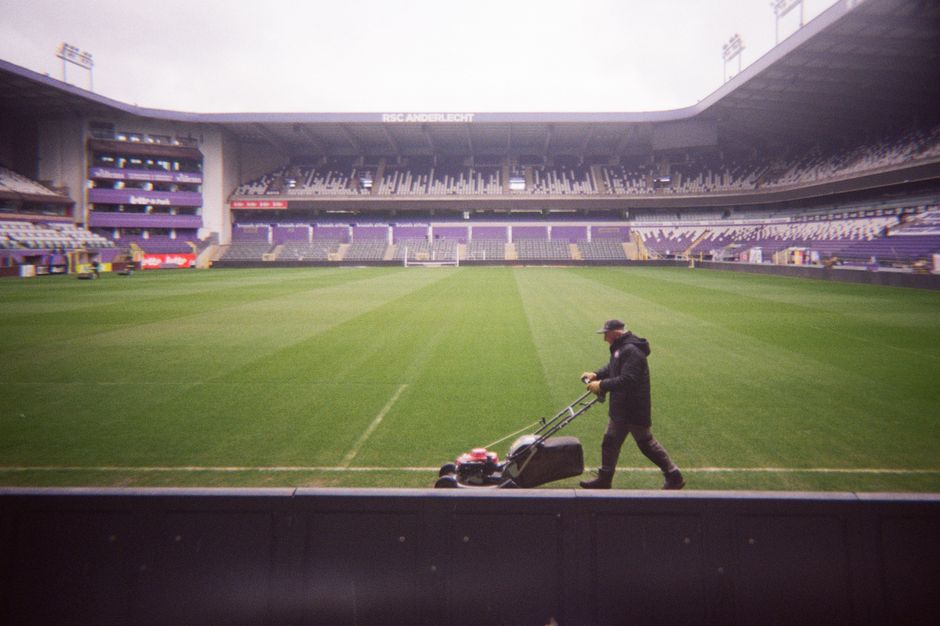 A wide view of a sports stadium featuring a well-maintained, lush green playing field with freshly mowed grass and visible mowing stripes. Surrounding the field are tiered seating areas with blue and 